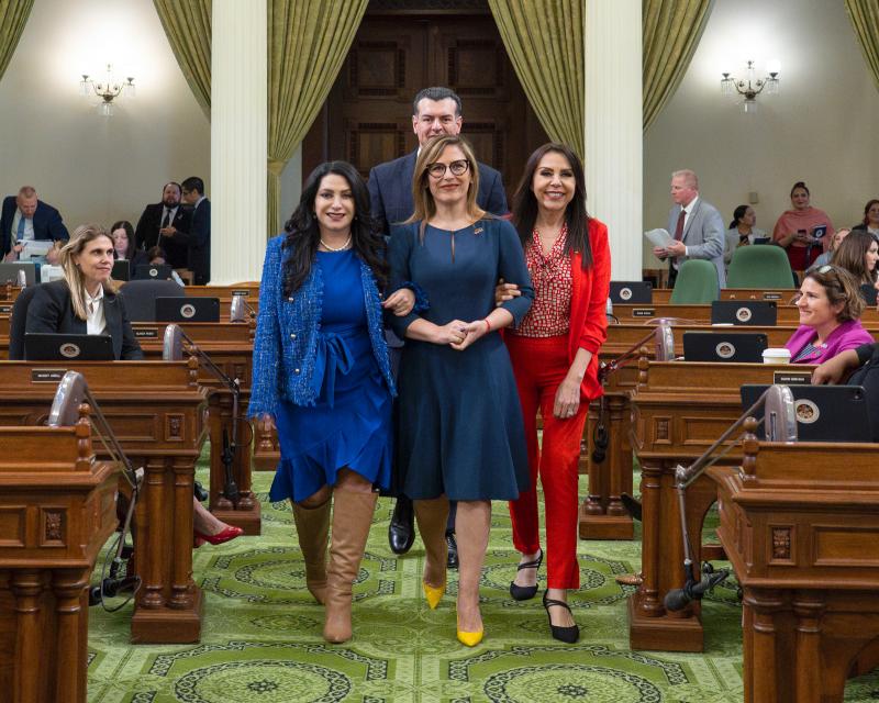 Senator Susan Rubio, actress Angélica Vale, and Assemblymember Blanca Rubio walk arm in arm down the center aisle of the California State Assembly chamber. Assemblymember Avelino Valencia is visible behind them. The group is smiling and surrounded by other legislators seated at their desks, with green carpet and tall columns framing the scene.