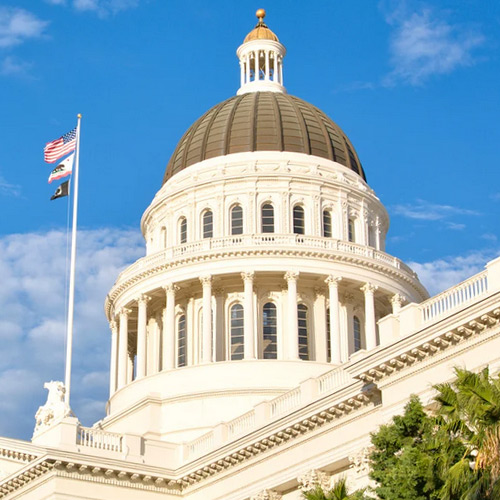 Close up shot of the California Capitol dome over a blue sky. The American flag flies on the flag pole above the California and POW flags