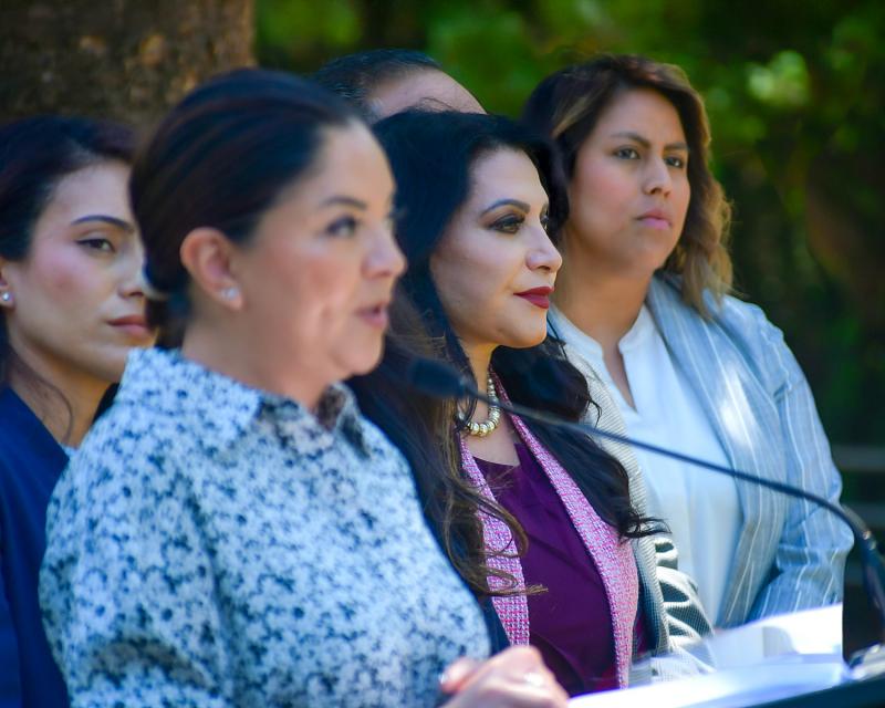 Four women legislators stand at a podium at the State Capitol during a press conference, with Senator Lena Gonzalez speaking while the others look on. Trees provide a green backdrop.