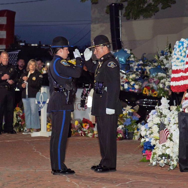 Two uniformed honor-guard officers exchange a salute during an evening memorial service, surrounded by wreaths, flowers, and small U.S. flags