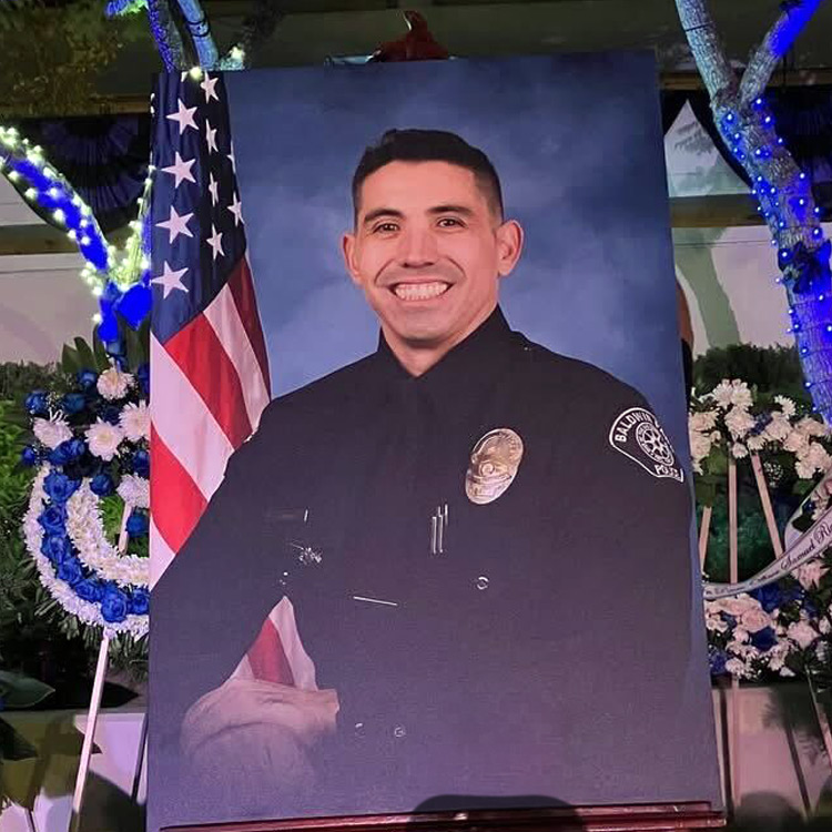 Large portrait of smiling Baldwin Park Police Officer Samuel Riveros in dress uniform, displayed beside an American flag and blue-white floral wreaths at a memorial