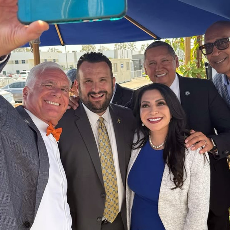 Senator Susan Rubio smiles with a group of officials and community leaders under a blue umbrella at a Foothill Gold Line event. The group is gathered for a selfie, dressed in business attire, with several people placing their arms around each other in celebration.
