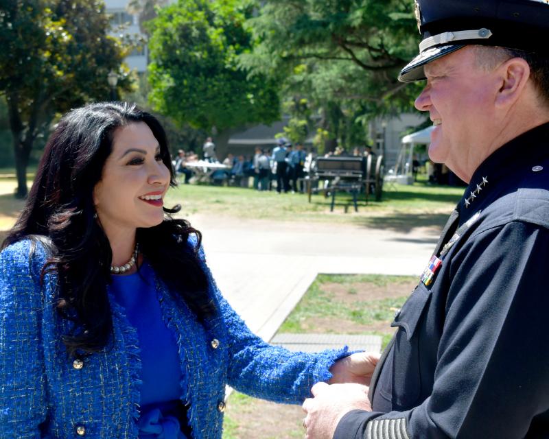 Senator Susan Rubio meets with LAPD Chief Jim McDonnell on the State Capitol grounds. They are outdoors on a sunny day, surrounded by trees, with a group of uniformed individuals gathered in the background.