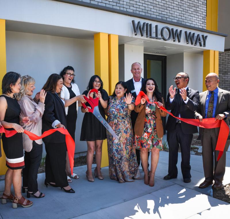 Senator Susan Rubio joins community leaders celebrating the Willow Way grand opening with a ribbon cutting. A woman in black holds oversized scissors while the group cheers